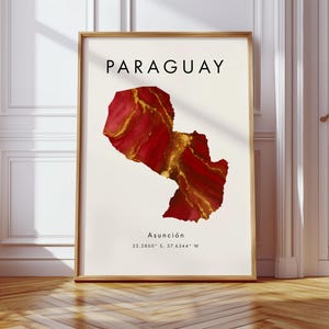 May include: A framed print featuring the map of Paraguay in shades of red and gold. The word "PARAGUAY" is printed at the top, with "Asunción" and coordinates below. The artwork is displayed in a room with a wooden floor and white walls.