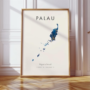 May include: A framed print showing a map of Palau in blue and gold on a cream backdrop. The word "PALAU" is printed in black at the top, with "Ngerulmud" and coordinates below. The frame is light brown. The print is displayed on a wooden floor.