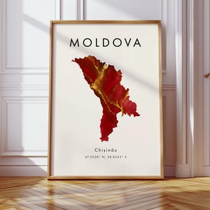 May include: A framed print showing a map of Moldova in red and gold hues. The word "MOLDOVA" is printed in black at the top, with "Chișinău" and coordinates below. The artwork is displayed in a room with a wooden floor.