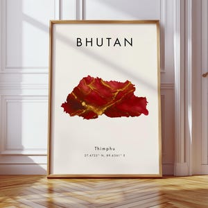 May include: A framed art print with the word "BHUTAN" above a red and gold abstract map of Bhutan. Below the map, the text reads "Thimphu" and coordinates. The print is in a wooden frame, displayed on a wooden floor.