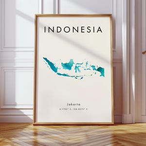 May include: A framed print with the word "INDONESIA" in black capital letters above a teal watercolour map of the country. Below the map, the city "Jakarta" and coordinates are printed in black. The frame is a light brown colour.