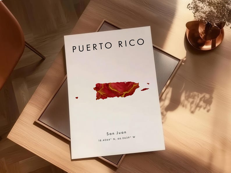 May include: A white print with the words "PUERTO RICO" in black, and a red and gold map of Puerto Rico. The print also includes the text "San Juan" and coordinates. The print is displayed on a wooden table.