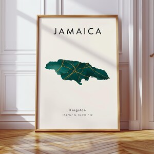May include: A framed art print with the word "JAMAICA" at the top. Below is a teal and gold map of Jamaica. The city "Kingston" and coordinates are printed below the map. The frame is a light brown colour.