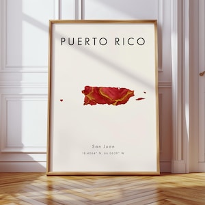 May include: A framed art print with the text "PUERTO RICO" above a red and gold map of Puerto Rico. The city "San Juan" and coordinates are printed below the map. The frame is light brown. The print is displayed on a wooden floor.
