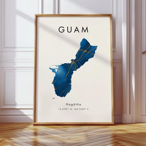 May include: A framed print featuring a map of Guam in shades of blue and gold. The word "GUAM" is printed at the top, with "Hagåtña" and coordinates below. The artwork is displayed in a room with a wooden floor and white walls.