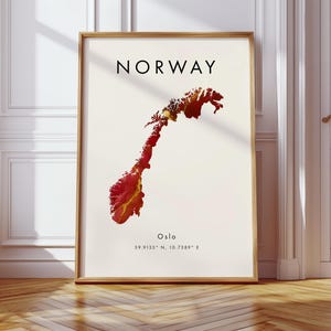 May include: A framed print showing a map of Norway in red and yellow hues. The word "NORWAY" is printed in black at the top, with "Oslo" and coordinates below the map. The frame is a light brown colour.