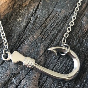 May include: A silver necklace with a hook pendant. The hook is a simple design with a rounded end. The necklace is on a wooden surface.