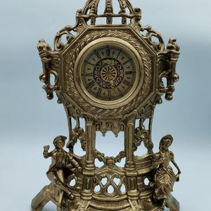 Op de afbeelding: Omslachtige goudkleurige mantelklok met een ronde wijzerplaat met Romeinse cijfers. De klok heeft een decoratief frame met figuren van mensen en een uitgebreide bovenkant. De klok is ongeveer 30 cm hoog.