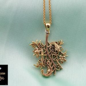 Puede incluir: Collar con colgante dorado con un diseño detallado y ornamentado. El colgante presenta una espada, llaves, flores y una luna creciente. El collar cuelga de una delicada cadena dorada sobre un fondo verde azulado suave.