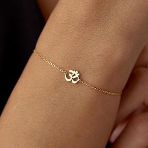 以下が含まれることがあります： 繊細なゴールドカラーのブレスレットで、Omのシンボルが特徴です。ブレスレットは、Omのシンボルをチャームとして使用した細いチェーンです。ブレスレットは手首に着用されており、背景は暗い色です。