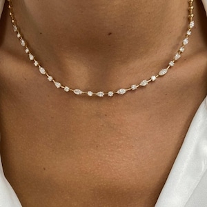Puede incluir: Un delicado collar de oro con pequeñas gemas brillantes. El collar tiene una fina cadena con piedras redondas y en forma de marquesa alternadas, creando un diseño refinado y elegante. El collar se lleva alrededor del cuello.