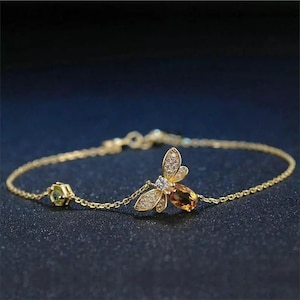 May include: A delicate gold bracelet featuring a bee pendant with sparkling accents. The bee has a central amber-colored stone and is adorned with small, clear crystals. A small, green-toned stone is set on the bracelet chain.