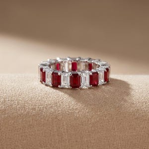 Può includere: Un anello a fascia eternity con un'alternanza di rubini rossi e diamanti bianchi trasparenti, entrambi con taglio a smeraldo e incastonati in una banda di metallo color argento. Le pietre formano un motivo continuo lungo tutto l'anello.