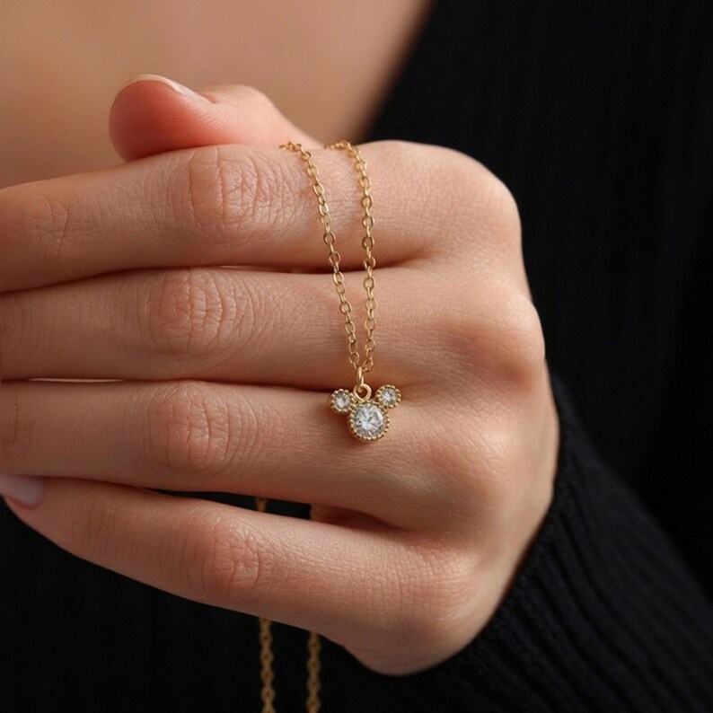 May include: A gold-colored necklace with a pendant shaped like a mouse head. The pendant features a large, round, clear gemstone in the center, with smaller gemstones on either side. The necklace is held in a hand against a dark background.