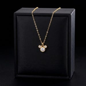 May include: A gold-colored necklace with a pendant shaped like a mouse head. The pendant features a large, round, clear gemstone in the center, with smaller gemstones on either side. The necklace is displayed on a black box.