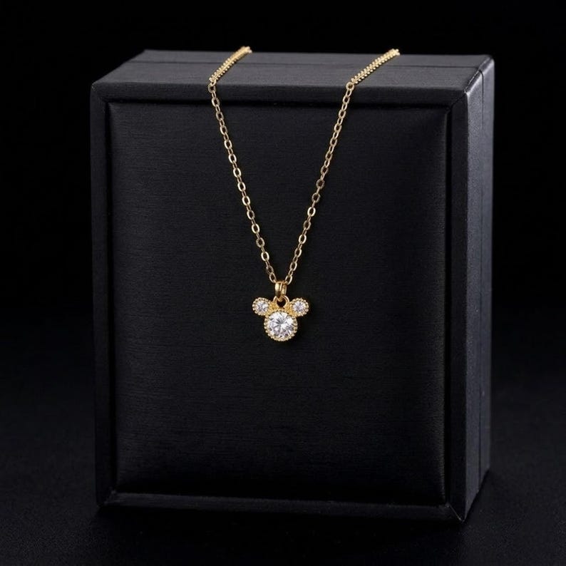 May include: A gold-colored necklace with a pendant shaped like a mouse head. The pendant features a large, round, clear gemstone in the center, with smaller gemstones on either side. The necklace is displayed on a black box.