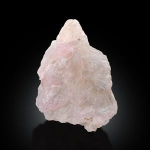 Puede incluir: Un espécimen de cristal de cuarzo rosa en bruto y sin cortar. La piedra tiene un tono rosa pálido y una superficie rugosa y texturizada. El cristal se presenta sobre un fondo negro, resaltando su belleza natural. Ideal para coleccionistas de cristales.