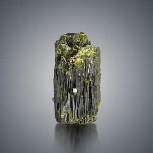 Könnte beinhalten: Ein rohes, natürliches Mineralexemplar mit einem dunklen, gestreiften Körper und leuchtend grünen und goldenen Akzenten. Die raue Oberfläche des Minerals reflektiert das Licht und zeigt seine kristalline Struktur. Das Mineral ist vor einem grauen Hintergrund platziert.