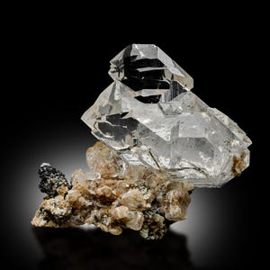May include: A clear quartz crystal formation with a large, faceted crystal on top. The crystal sits on a base of smaller, tan-colored crystals and a dark, black mineral. The background is black.