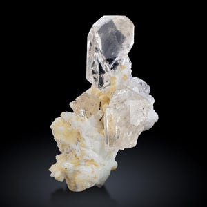 May include: A clear, crystalline mineral specimen with a large, upright crystal formation. The main crystal is transparent, with a complex structure. The base is a mix of white and yellow-toned minerals, set against a black background.