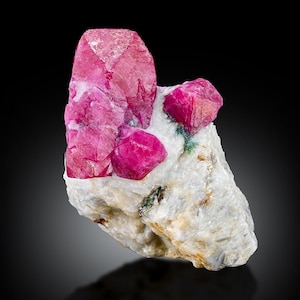 Op de afbeelding: Een ruw mineraal exemplaar met levendige, dieproze kristalstructuren ingebed in een ruwe, gebroken witte matrix. Het cluster vertoont scherpe, geometrische facetten op de magentakleurige kristallen, die contrasteren met de getextureerde, ondoorzichtige basis.