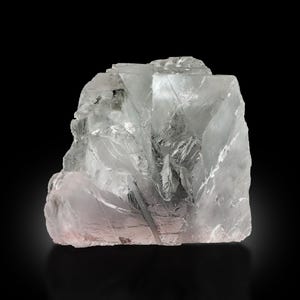 Könnte beinhalten: Ein durchscheinendes, kristallines Mineralexemplar mit rauer, eckiger Form. Das Mineral hat einen blass, eisigen blau-grauen Farbton mit subtilen rosa Untertönen. Die Oberfläche reflektiert das Licht und erzeugt einen schimmernden Effekt vor dem schwarzen Hintergrund.