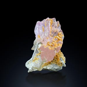May include: A natural mineral specimen featuring a cluster of translucent, light purple crystals with a golden-yellow, textured matrix. The base is a mix of clear and pinkish hues, set against a black background.