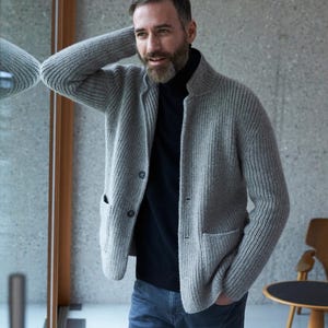 May include: A light gray, ribbed-knit cardigan with two front pockets and button closures. The cardigan is worn over a black turtleneck and paired with blue jeans. The style is casual and comfortable.