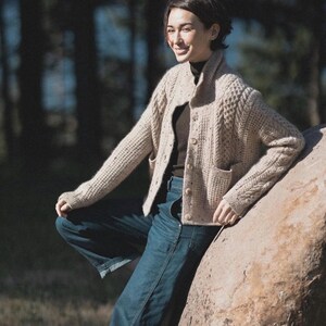 May include: A beige cable-knit cardigan with a button-up front and two front pockets. The cardigan is worn with a dark brown turtleneck and blue denim jeans. The person is leaning against a large rock in an outdoor setting.