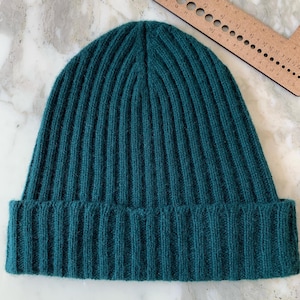 May include: Teal ribbed knit beanie hat. The hat is made of a soft, textured material and features a folded brim. The hat is a solid teal color and is suitable for cold weather.