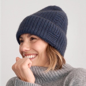 Könnte beinhalten: Eine marineblaue Strickmütze. Die Mütze besteht aus einem weichen Rippstrickmaterial. Die Person auf dem Bild trägt einen grauen Rollkragenpullover. Der Hintergrund ist einfarbig weiß.