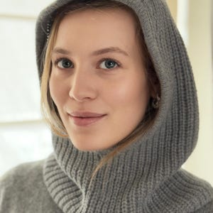 May include: A grey knitted hood and cowl. The hood is up, framing the face. The cowl is ribbed and sits around the neck. The knitwear looks soft and comfortable, suitable for cold weather. The overall style is casual and practical.