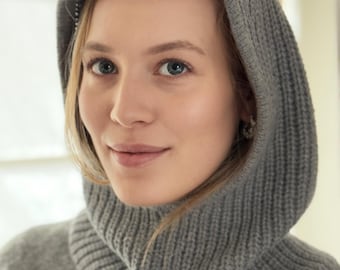 May include: A gray knit hood and cowl combination. The hood is pulled up, framing the face. The cowl is ribbed and sits around the neck. The knitwear appears soft and comfortable, suitable for cold weather. The overall style is casual and practical.
