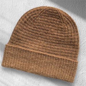 Può includere: Un berretto lavorato a maglia a coste marrone. Il cappello ha un bordo ripiegato e un design aderente. Il colore è un marrone caldo e terroso e il motivo a maglia fornisce consistenza. Questo cappello invernale è adatto al freddo.