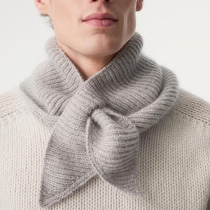 May include: A light gray knitted scarf tied around the neck. The scarf is made of a soft, ribbed material and is worn over a cream-colored sweater. The scarf's ends form a pointed shape.