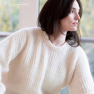 May include: Cream-colored knit sweater with a ribbed texture. The sweater has a crew neck and a boxy design. The image also includes text that reads "SANDEL PULLOVER" and "DESIGNED BY SASCHA WOBIDO" with a "SHOP PATTERN" button.