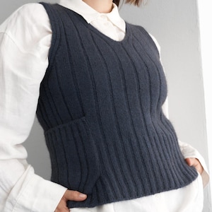 Può includere: Un gilet lavorato a maglia blu scuro con scollo a V e trama a coste. Il gilet ha una piccola tasca sul lato sinistro e viene indossato sopra una camicia bianca con colletto. Il colore del gilet è un blu intenso e smorzato.