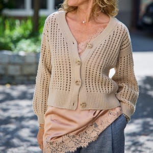 Könnte beinhalten: Eine beige Strickjacke mit grobem Lochmuster, V-Ausschnitt und vier Knöpfen, getragen über einem pfirsichfarbenen Satin-Top mit zarter Spitzenborte am Saum, kombiniert mit einer dunkelgrauen Hose mit sichtbarer Textur und lockerer Passform.