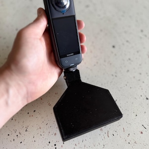 May include: A black Insta360 camera with a screen and a wide-angle lens, connected to a black, angled base. The camera is held in a hand against a speckled background. The Insta360 logo is visible on the screen.