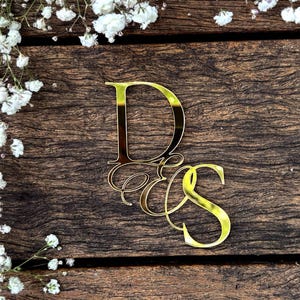 May include: A gold-coloured monogram cake topper with the letters "D", "E", and "S" in a stylish script. The topper sits on a rustic wooden surface, with white flowers in the upper left corner. The overall effect is elegant.