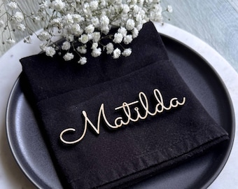 May include: A wooden name tag reading "Matilda" in cursive sits on a black napkin, on a black plate. White flowers are arranged above the napkin. The image suggests a place setting for a special occasion or event.