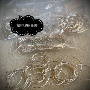 Op de afbeelding: Een close-up van een stapel zilveren metalen wijncharms. De charms zijn verpakt in doorzichtige plastic zakken met de tekst "Wine Charm Hoops" op een zwart wolkvormig label.