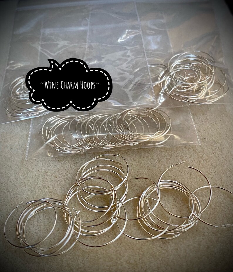 Op de afbeelding: Een close-up van een stapel zilveren metalen wijncharms. De charms zijn verpakt in doorzichtige plastic zakken met de tekst "Wine Charm Hoops" op een zwart wolkvormig label.