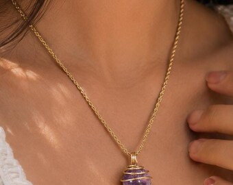 Puede incluir: Un collar de cadena de cuerda dorada con un colgante compuesto por una piedra preciosa de amatista morada esférica y de corte rugoso, encerrada en una jaula de alambre decorativa de tono dorado. El colgante cuelga de una argolla dorada.