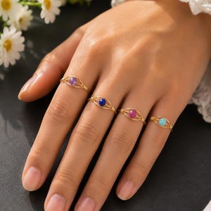 May include: Four gold-toned wire-wrapped rings, each featuring a single small round gemstone. The stones are purple, dark blue, pink, and light blue. The wire design forms an oval shape around each bead, with coiled wire details on the sides.