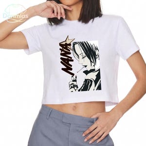 May include: White crop top with a black and white anime illustration of a person smoking, and the word "NANA" in black. The design includes a star-shaped element. The shirt is a casual, short-sleeved style.
