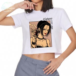 Puede incluir: Top corto blanco con una ilustraci&oacute;n de estilo manga de una mujer con cabello negro, labios rojos y un cigarrillo. El gr&aacute;fico est&aacute; enmarcado en un cuadro marr&oacute;n claro, con el texto "#Sixfanarts" y "OSAKI NANA" impreso en la camiseta.
