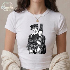 May include: White t-shirt featuring a black and white anime-style illustration of a person with a guitar. The person is wearing a black hat, jacket, and plaid skirt. The shirt has the text "Donmlas" in the upper left corner.