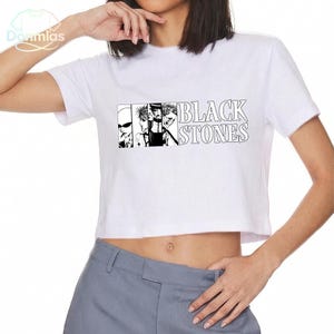 May include: White crop top with a black and white graphic of four figures and the text "BLACK STONES". The shirt has short sleeves and a crew neck. The graphic is in a rectangular box.