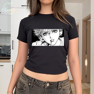 May include: Black crop top featuring a black and white manga illustration. The graphic depicts a person with spiky hair, multiple piercings, and a chain necklace. The top has a crew neck and short sleeves.